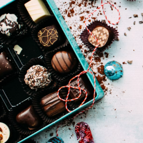 An aquamarine coloured box on chocolates on a white counter filled with enrobed chocolates with some outside the box artistically placed. Image by the Foodess on Unsplash