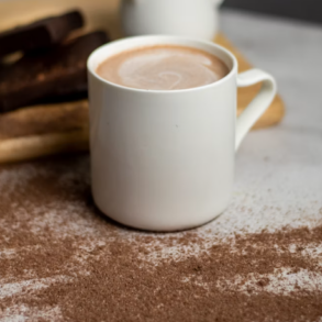 A white mug of hot chocolate with a frothy top and with cocoa powder covering the counter infront of it. Image by Chester Toh on Unsplash